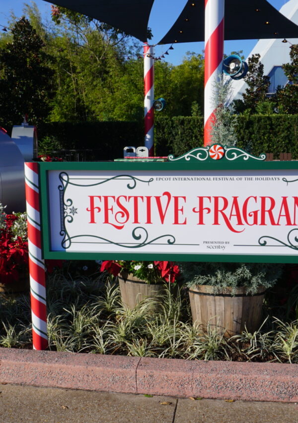A holiday display at EPCOT featuring a large sign that reads “Festive Fragrances” presented by Scentsy. The scene includes candy-cane–striped poles, poinsettias, greenery, and a giant ornament-shaped showcase filled with festive Scentsy products.