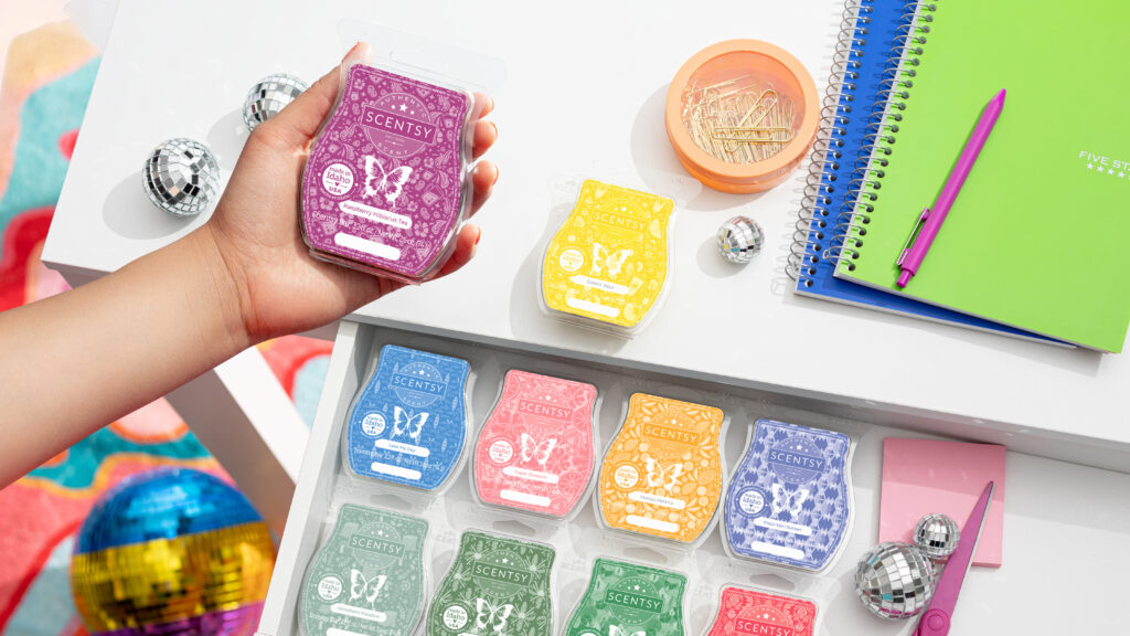 A hand holds a Scentsy Wax bar over a desk and drawer filled with Scentsy wax bars, notebooks, pens and other office supplies.