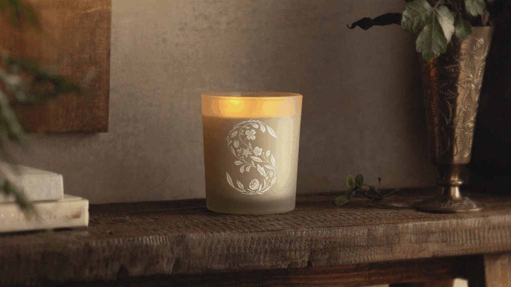 A glowing scented candle baring the Scentsy S logo sits on a dark wood mantle. Books, a painting, and a brass vase can be seen on the perifery. 