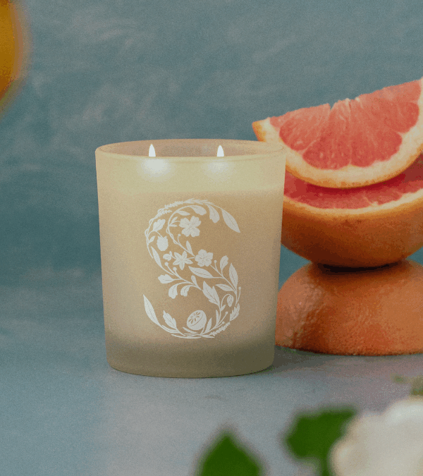 A Scentsy Blue Grotto Premium Scented Candle sits beside sliced blood oranges, with flower blossoms and lemons dangling from branches surrounding it.