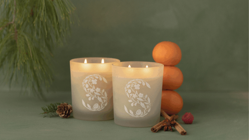 Two glowing scented candles baring the Scentsy S logo sit on a dark green backdrop. Oranges, cinnamon sticks, a pinecone, a raspberry, and pine boughs surround the candles to denote the fragrances notes of the scented wax. 