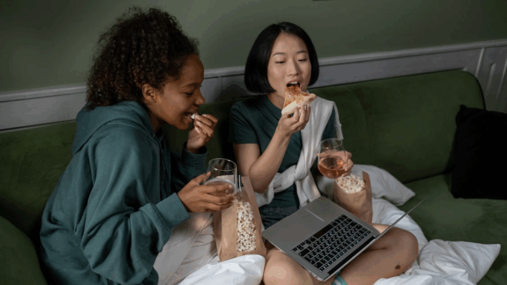 Two female friends sit and watch movies on a laptop while eating girl dinner. 