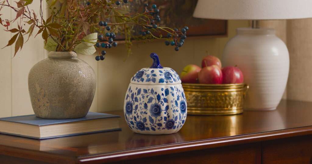 A Scentsy Indigo Vine Pumpkin Warmer is displayed on an entry table next to a lamp, bowl of apples and fall decor. Wax melts in this warmer create cozy fall scents.