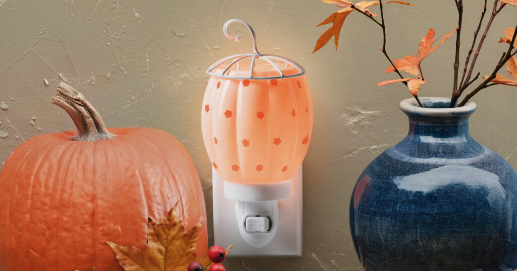 A Pumpkin Posy Mini Warmer plugged into a wall near a pumpkin and fall leaves, ideal for warming wax melts in cozy fall fragrances.